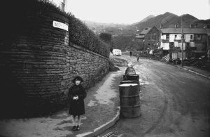A young survivor stands at the corner of Moy Road; behind her, a scene of complete ruin.